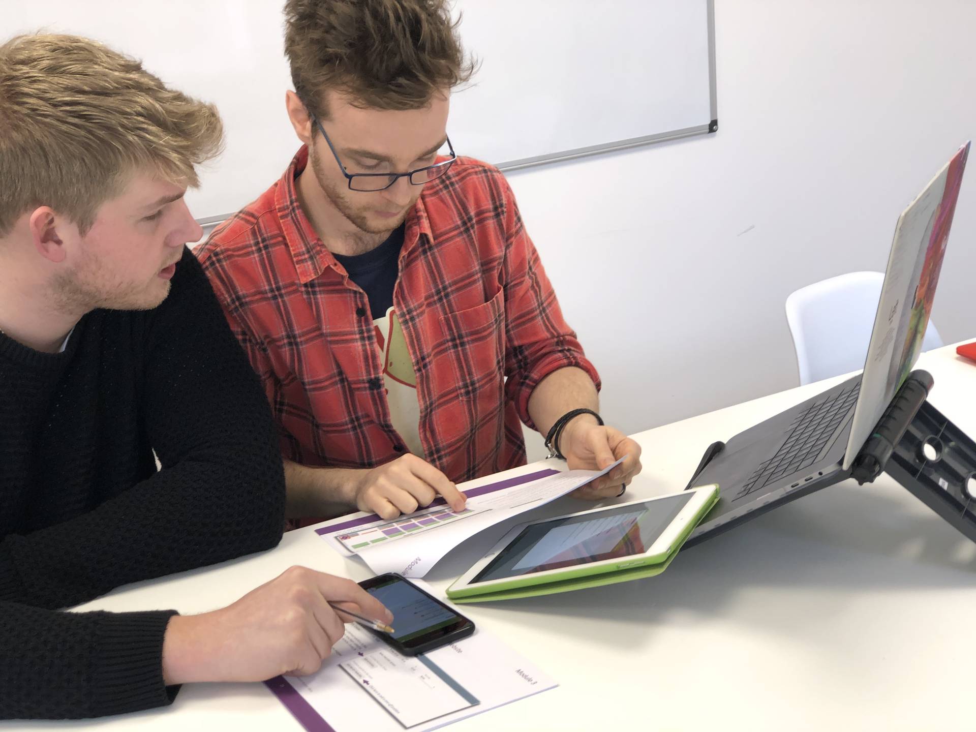 Two men are seated at a white table, appearing to collaborate on a task involving multiple digital devices and physical documents. One man points at a smartphone, while the other reviews papers next to a laptop and tablet.