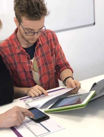 Two men are seated at a white table, appearing to collaborate on a task involving multiple digital devices and physical documents. One man points at a smartphone, while the other reviews papers next to a laptop and tablet.