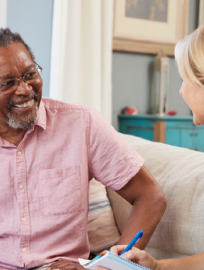 A man with greying hair and a beard, wearing glasses and a pink shirt, sits on a light-coloured sofa, smiling and looking at a blonde woman in a navy shirt who is sitting opposite him, writing on a notepad. They appear to be engaged in a conversation in a comfortable home setting.